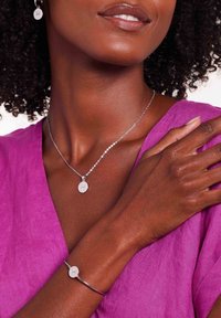 Zilveren sierraden omvatten een delicate kettinghalsketting en armband met ronde bedels. De outfit is van levendig paarse stof met een gladde textuur.