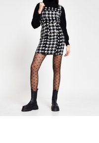 Schwarzes und weißes Houndstooth-Kleid mit goldenen Knöpfen, kombiniert mit schwarzen gepunkteten Strumpfhosen und klobigen schwarzen Knöchelstiefeln. Glatte Textur und taillierte Passform.