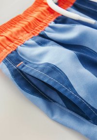 Costumi da bagno blu con motivi ondulati, vita elastica arancione, lacci bianchi e dettagli di cucitura visibili. Realizzati in tessuto leggero.