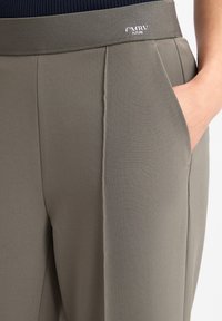 Grijze broek met een gladde textuur, voorzien van een brede tailleband, zijzakken en subtiele plooien. Logo detail op de tailleband.