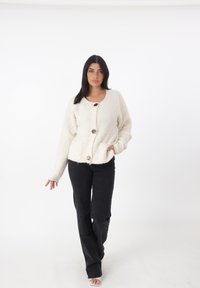 Cardigan crema e morbido con scollatura rotonda, tre bottoni e due tasche frontali, abbinato a pantaloni neri a zampa.