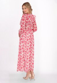 Maxikleid aus transparenter Stoff, mit einem pinken Blumenmuster, Rüschendetails an den Schultern und einer taillierten Passform. Kombiniert mit beigen Heels.