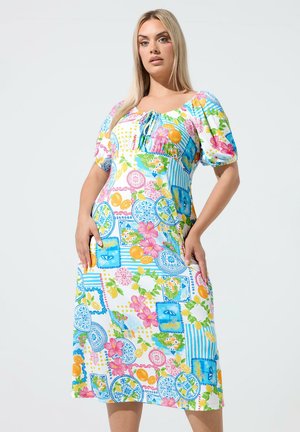 Vrouw met lang blond haar, gekleed in een kleurrijke, met patronen versierde midijurk met pofmouwen, staand tegen een effen witte achtergrond.