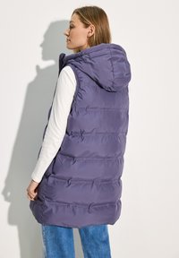 Gilet rembourré violet avec capuche, featuring un design matelassé, des coutures visibles et des poches latérales. Porté par-dessus une chemise à manches longues blanche.