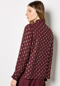 Burgundy blouse with gold leaf pattern, soft fabric, long sleeves with gathered cuffs, high ruffled collar, and a relaxed fit.