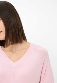 Lyserød, V-hals striktrøje med en blød tekstur. Har drop-skuldere og diskrete detaljer langs sømmene.