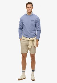 Sudadera azul claro con puños acanalados, pantalones cortos beige y suéter de punto en color crema atado en la cintura. Zapatillas blancas con detalles en beige.