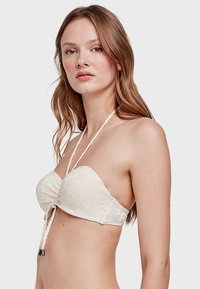 Strapless crèmekleurige bikini-top met een getextureerd spiraalpatroon, verstelbare bindstrikken en metalen accenten. Zacht materiaal met een gladde afwerking.