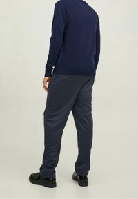 Uomo in piedi di spalle, che indossa un maglione blu navy, pantaloni blu scuro e scarpe nere, contro uno sfondo chiaro e semplice.
