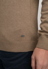 Lichtbruine trui met geribde manchetten, gladde textuur en een klein metallic logo aan de onderzijde, gecombineerd met donkere broek.