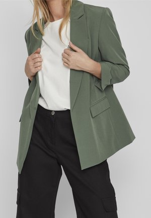Persoon draagt een groene blazer over een wit shirt met zwarte, hoog uitgesneden cargobroek, handen houden de revers van de blazer vast.