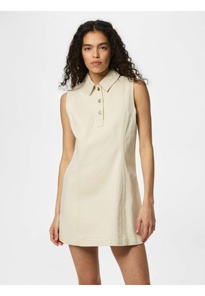 Kvinde med krøllet hår iført en ærmeløs, cremefarvet kjole med krave og knapdetaljer, stående foran en ensfarvet hvid baggrund.