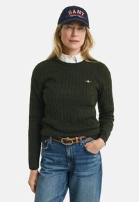 Dunkelgrüner Kabelstrickpullover kombiniert mit einem weißen Hemd und Jeans. Die Accessoires umfassen eine marineblaue Kappe mit "GANT"-Logo und einen braunen Gürtel.