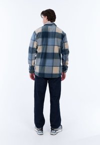 Man met zijn rug naar de camera, gekleed in een groot geruit blauw en beige jack, donkere broek en grijze sportschoenen, voor een witte achtergrond.