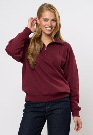 Burgundy kvartshals sweatshirt tillverkad av mjukt material, med ribbade muddar och en liten logotyp på bröstet, i kombination med mörka jeans.