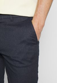 En mans högra hand vilar i fickan på mörka texturerade byxor, iklädd en ljusbeige skjorta som är instoppad.