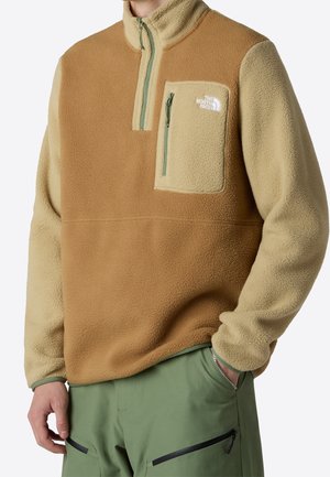 Homme portant un pull polaire beige et crème avec une poche poitrine zippée et un pantalon vert avec poches zippées, debout avec une main dans la poche.