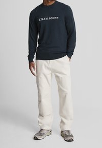 Maglione a girocollo lavorato a maglia di colore blu navy con la scritta bianca "LYLE & SCOTT", pantaloni cream a gamba larga e sneaker grigie con un motivo decorativo.