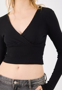 Woman wearing a black ribbed long-sleeve cropped top and grey high-waisted jeans, showing midriff against a white background.