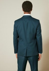 Mörk teal kostym med en enda slits bak, skräddarsydd passform och matchande byxor, med knappar på ärmarna och en vit skjorta under.
