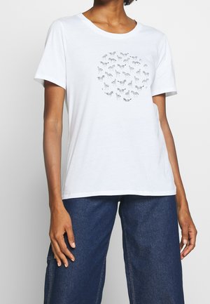 Nainen, joka pukeutunut valkoiseen T-paitaan, jossa on mustavalkoinen pyöreä seepra-kuvio, yhdistettynä tummansinisiin farkkuihin.