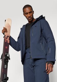 Giacca da sci blu navy con cappuccio, dotata di tasche con zip e tessuto testurizzato. Tiene una tavola da snowboard con attacchi, indossata sopra una maglietta nera.