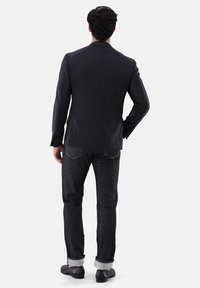 Dunkelgrauer Blazer mit schmaler Passform, kombiniert mit dunklen Jeans, hochgekrempelten Ärmeln und schwarzen Schuhen, von hinten vor einem weißen Hintergrund betrachtet.