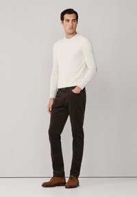 Hombre de pie con la mano en el bolsillo, vistiendo un suéter blanco de cuello redondo, pantalones marrón oscuro y zapatos de cordones marrón claro, frente a un fondo liso.