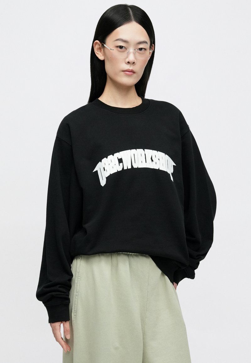 Zwarte sweatshirt met witte tekst "WORKSHOP" in een vet, gebogen lettertype. Heeft lange mouwen en een relaxte pasvorm. Gecombineerd met lichtgroene broeken.