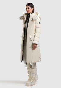 Beige puffer jas met een lange uitstraling, zijzakken, een met bont gevoerde capuchon en een zwarte rits. Combinatie met bijpassende geïsoleerde laarzen.