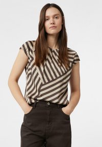 Frau mit langen braunen Haaren, die eine beige-braun gestreifte Kurzarmbluse und dunkle Hosen trägt, steht mit den Händen in den Taschen.