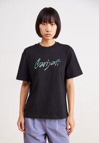 Camiseta de algodón negra con un logo verde brillante de "Carhartt" en el pecho. Corte relajado con mangas cortas. El modelo lleva pantalones de color púrpura claro.