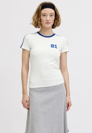 Mujer joven con cabello corto que lleva una camiseta blanca con ribete azul y número 81, falda gris y una cinta negra en la cabeza sobre un fondo liso.