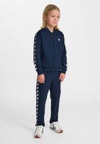 Tuta sportiva blu navy con strisce a zig-zag bianche sulle maniche e sui pantaloni. La giacca presenta una zip e un logo sul petto. Il modello indossa scarpe da ginnastica bianche e verdi.