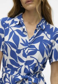 Camicia a maniche corte bianca con un motivo di foglie blu, dotata di colletto, chiusura a bottoni e dettaglio in vita annodato per un effetto aderente.