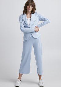 Costume bleu clair comprenant un blazer ajusté et un pantalon à jambes larges, fabriqué en tissu lisse. Porté avec un haut blanc et des baskets blanches.