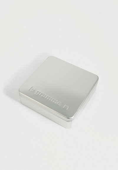 Taisnstūra formas sudraba metalizēta kārba ar noapaļotiem stūriem un uzrakstu "le gramme" uz vāka, novietota uz baltas virsmas.