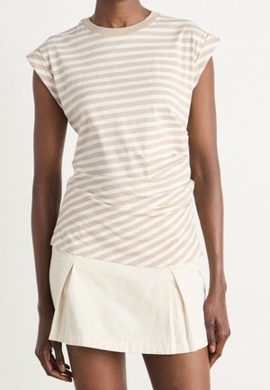 Person, die ein ärmelloses Top mit beige-weißen Streifen und einen weißen plissierten Minirock trägt, vor einem einfarbigen Hintergrund steht.