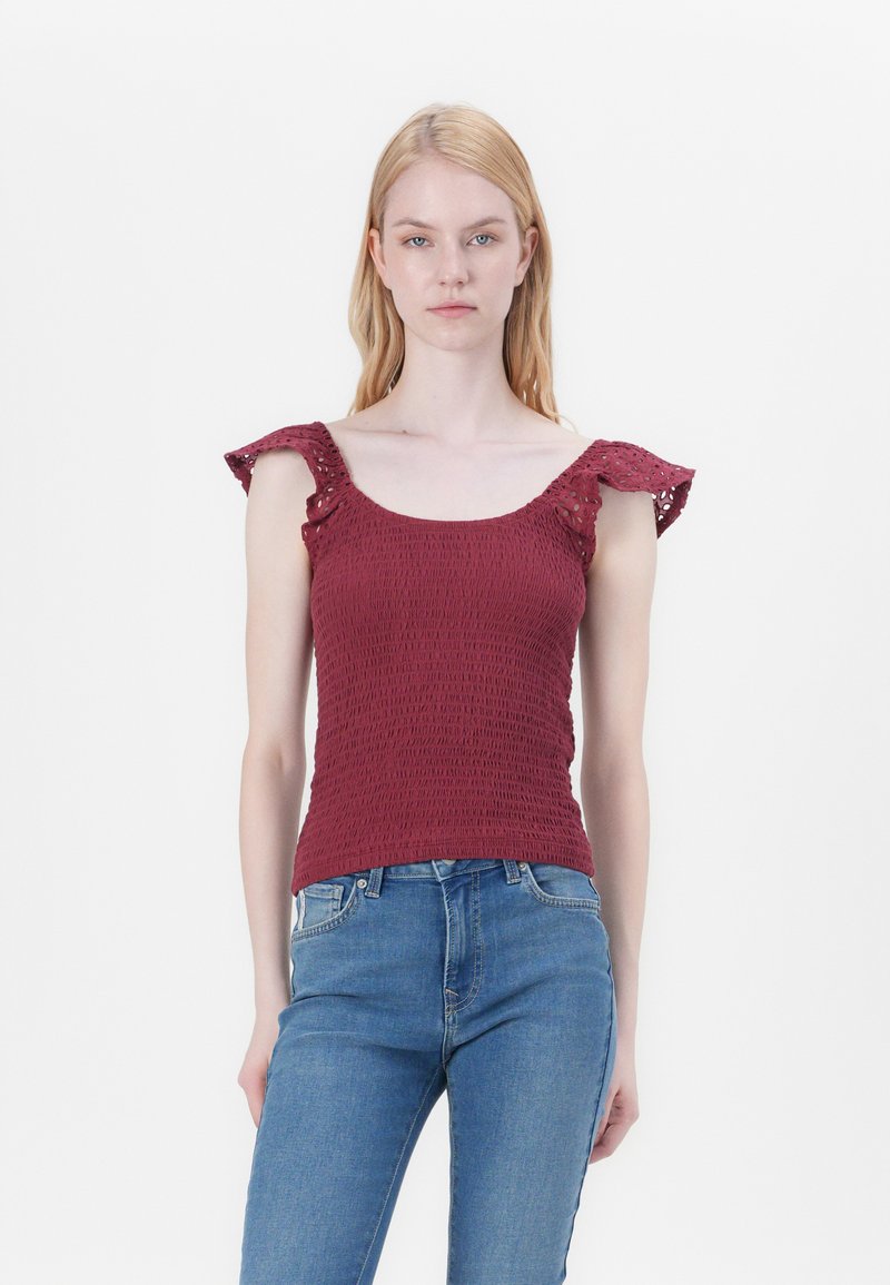 Burgundy smocked top with ruffled shoulder accents, made of textured fabric. Paired with light blue jeans, featuring a fitted design.