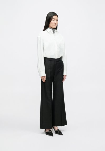 Witte overhemd met kraag en knopen, lange mouwen, gecombineerd met hooggetailleerde zwarte wijde broekspijpen en zwarte pumps met spitse neus.