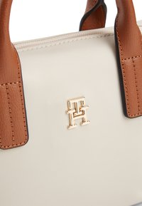 Κρεμ τσάντα χειρός με καφέ δερμάτινα χερούλια και χρυσό μεταλλικό διαπλεκόμενο λογότυπο "TH" μπροστά.