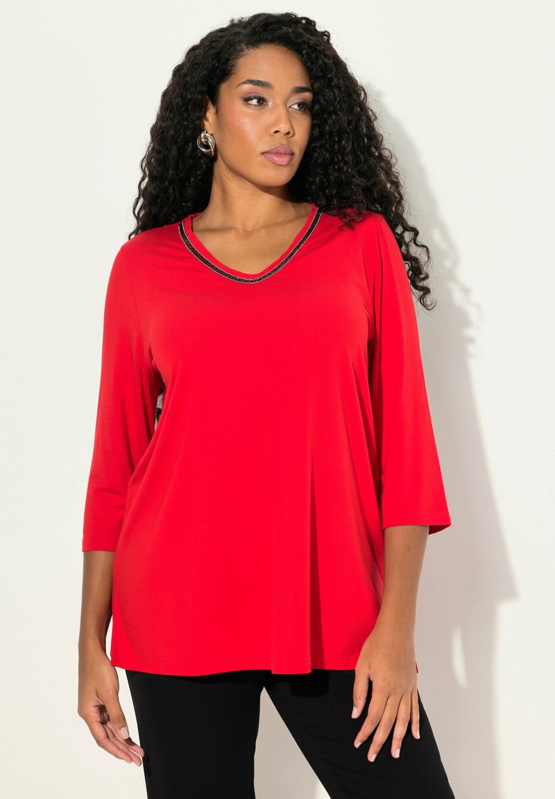 Červený top s dlouhými rukávy a výstřihem do V, obohacený černým akcentem kolem výstřihu. Vyrobený z měkké tkaniny, má volný střih, který jemně volně splývá.