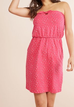 Femme portant une robe rose sans manches à petits pois blancs et taille froncée, debout devant un fond uni.