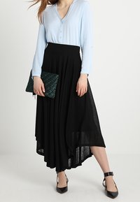Camicia azzurro chiaro con bottoni, gonna nera fluida asimmetrica, pochette verde trapuntata, tacchi neri. Tessuto morbido, trame sovrapposte, silhouette elegante.