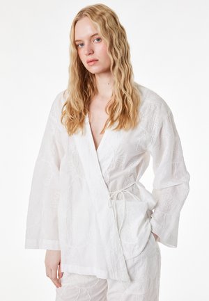 Blonde Frau mit welligem Haar, die eine weiße Wickelbluse und passende Hose mit bestickten Blumenmustern trägt, steht mit einer Hand in der Tasche.