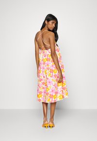 Vestido maxi floral multicolorido com padrões em rosa, amarelo e branco. Apresenta alças finas e um design com arco nas costas. Usado com sapatos de salto amarelos.