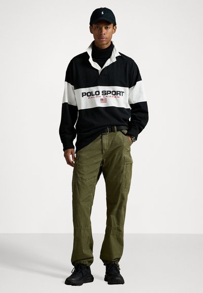 Zwart-wit oversized rugbyshirt met "POLO SPORT" tekst, olijfgroene cargobroek met zakken, zwarte sneakers en een zwarte pet.
