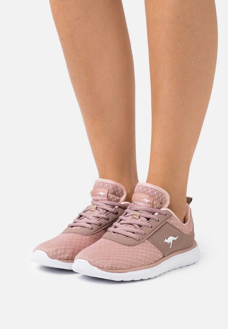 Personne portant des baskets KangaROOS en mesh rose avec des semelles blanches, debout sur une surface unie et claire.