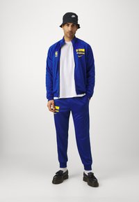 Mėlynas treningas su užtraukiamu švarku ir kelnėmis, kuriuose geltonos spalvos užrašai "Warriors" ir Nike logotipas. Juoda kibiro skrybėlė ir juodi batai užbaigia įvaizdį.