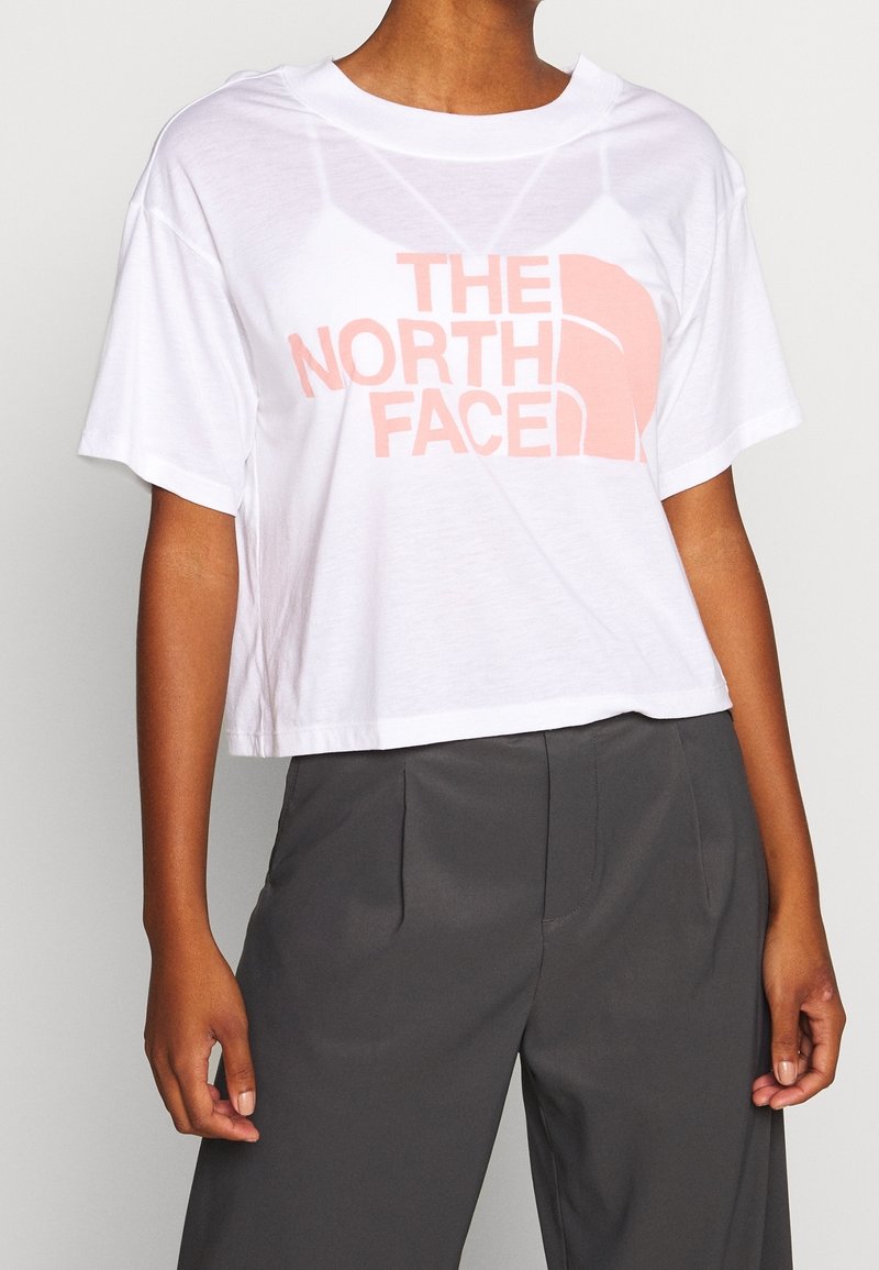 Bílý zkrácený tričko s lehce růžovým logem "THE NORTH FACE". Měkká látka, volný střih, kulatý výstřih a krátké rukávy. Žádné vzory ani akcenty.